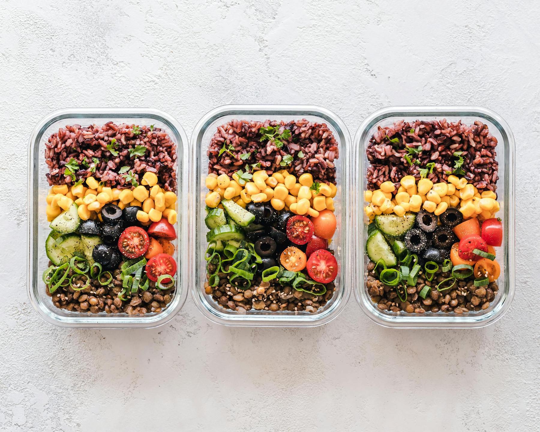 Three glass containers filled with layered ingredients: red rice, corn, tomatoes, cucumbers, olives, lentils, and scallions on a light background.