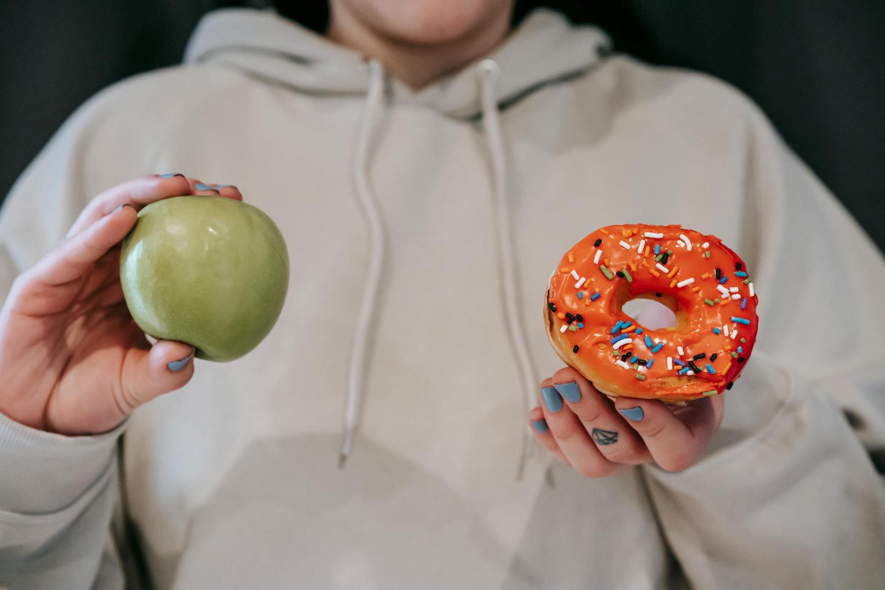 A person in a beige hoodie holds a green apple in one hand and an orange frosted donut with sprinkles in the other.