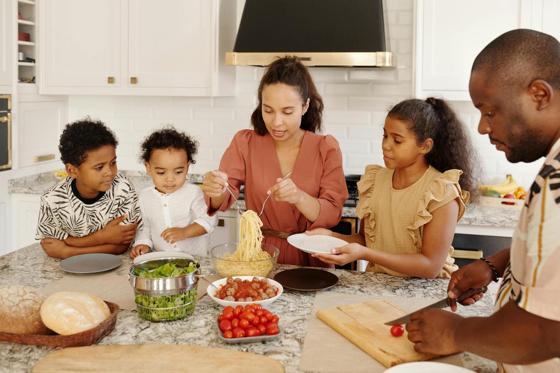 A family is gathered in a kitchen preparing a meal together, with pasta, vegetables, and bread on the counter.