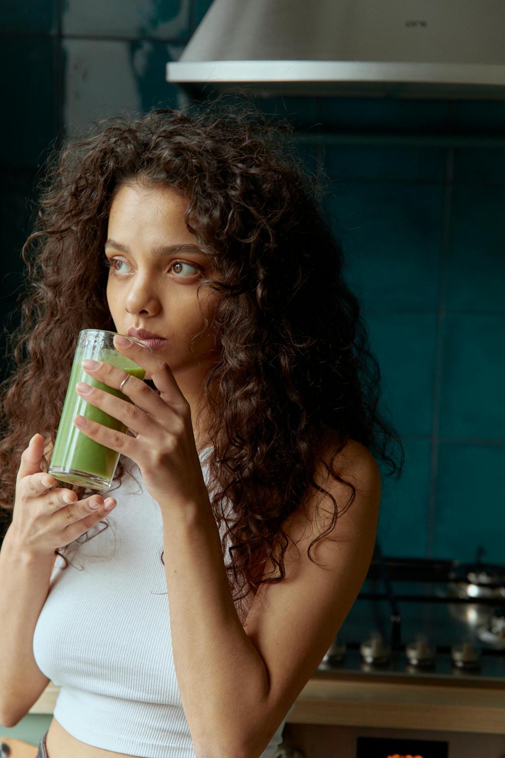A woman with curly hair in a white sleeveless top holds and drinks a green smoothie in a kitchen.