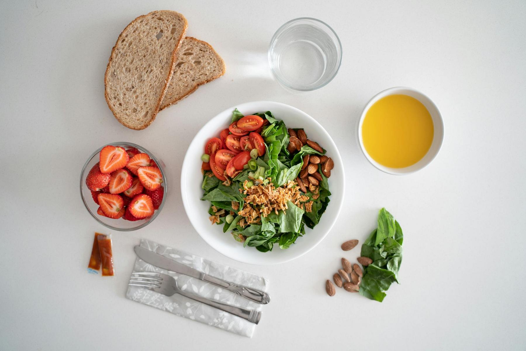 A meal with a salad bowl, sliced cherry tomatoes, almonds, two bread slices, a glass of water, a bowl of orange juice, a bowl of strawberries, cutlery, and sauce packets on a white table.
