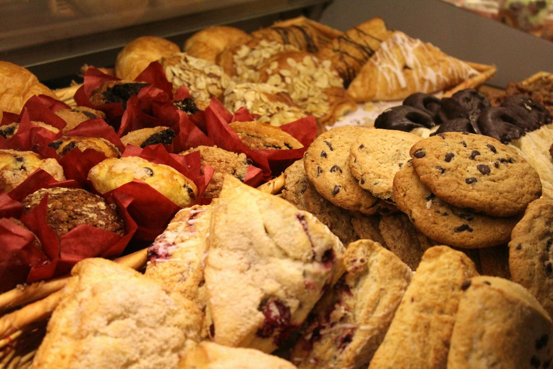 An assortment of baked goods including cookies, scones, muffins in red wrappers, almond pastries, and chocolate-covered treats displayed on a tray.