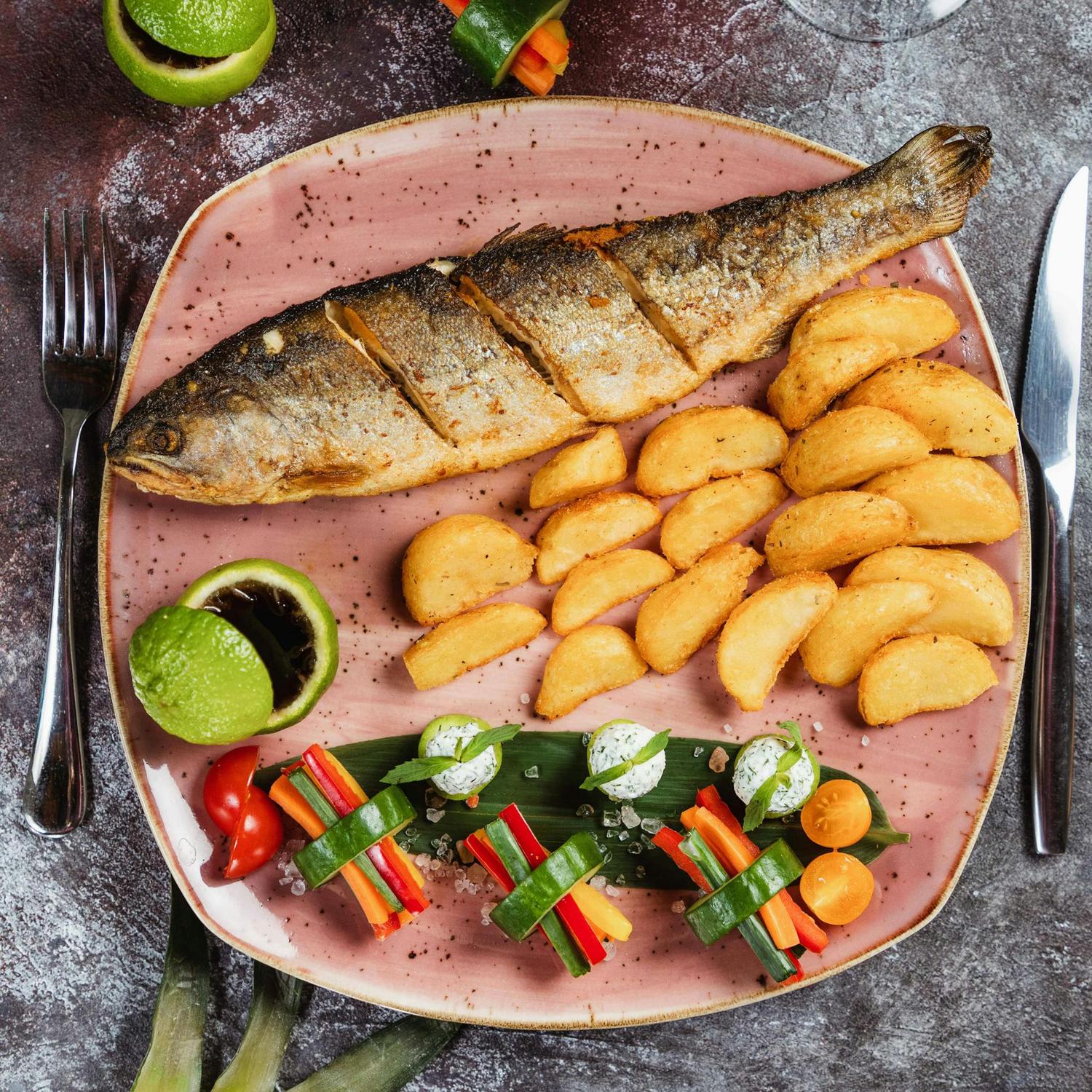 A whole roasted fish with crispy potato wedges, fresh lime halves, and assorted vegetable sticks with a creamy dip are served on a pink plate with a fork and knife beside it.
