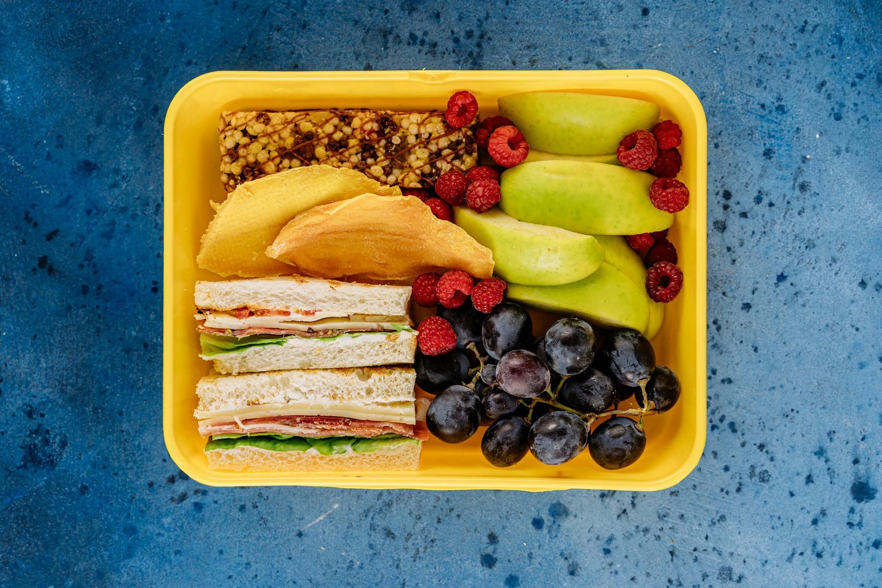 A yellow lunchbox containing sandwiches, green apple slices, grapes, raspberries, a granola bar, and pancakes on a blue background.