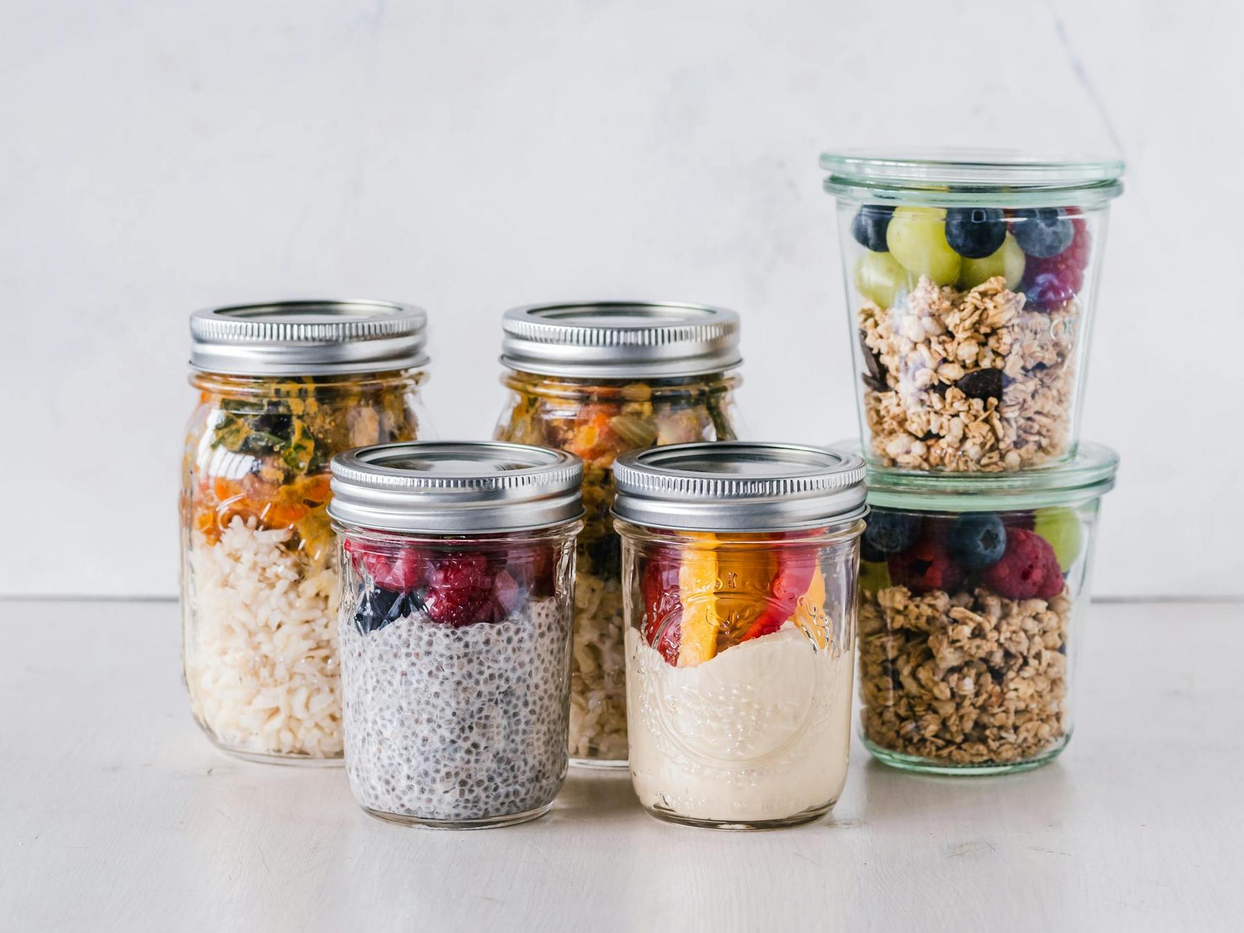 Six glass jars filled with items like rice with vegetables, chia pudding with berries, yogurt with fruit, and granola are arranged on a white surface.