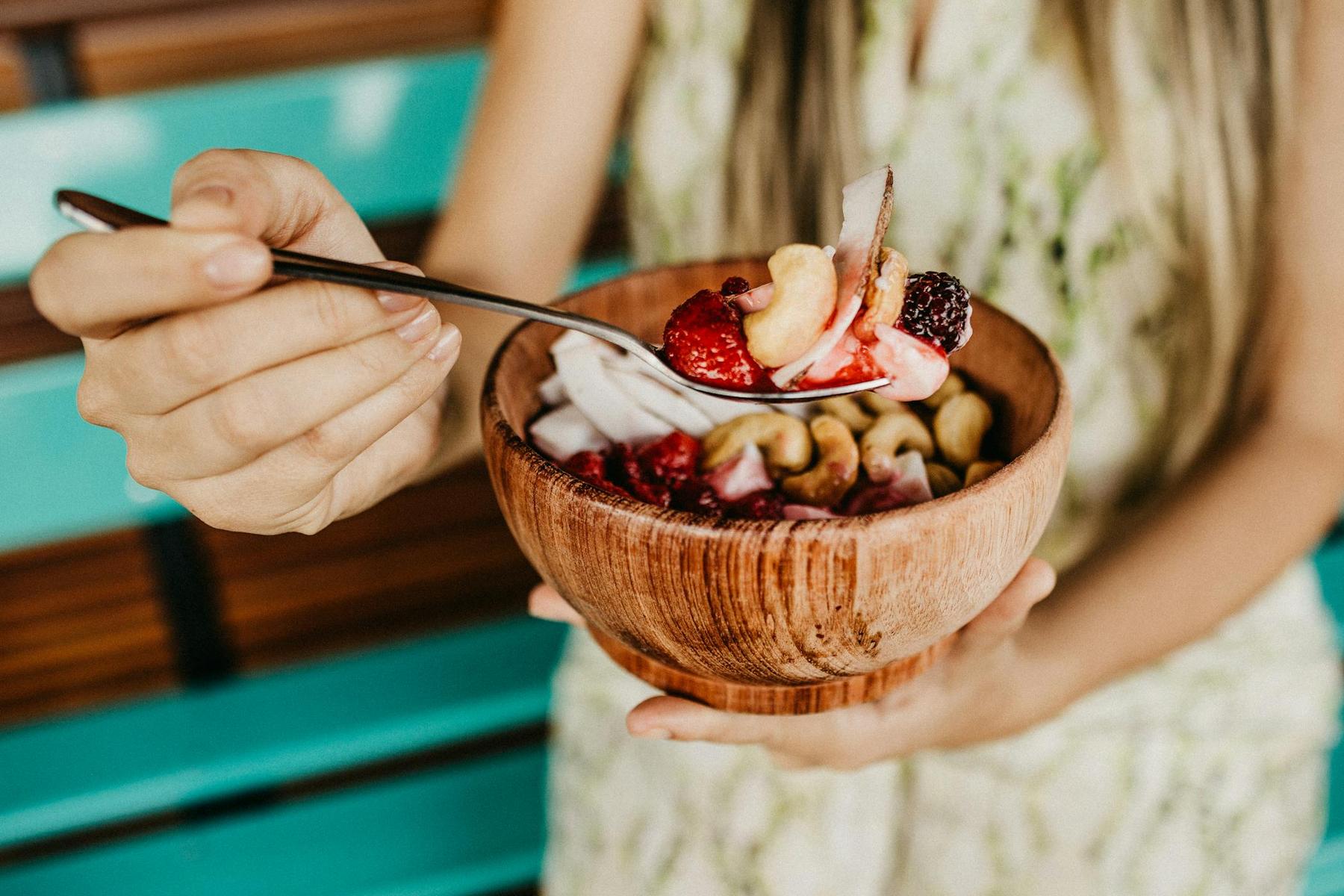 A person holding a wooden bowl filled with mixed fruits, nuts, and yogurt, using a spoon to scoop a bite. The background features turquoise and wooden elements.