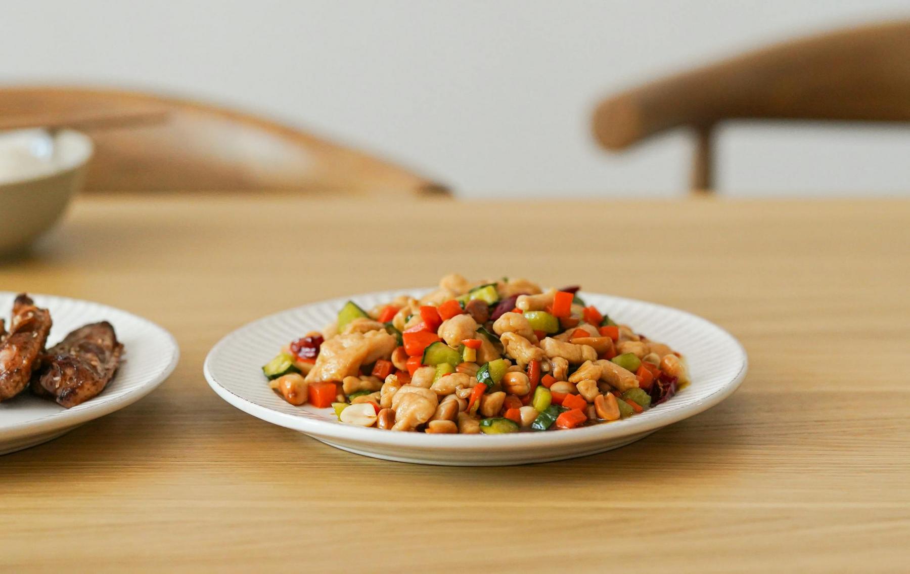 A plate of stir-fried chicken with diced vegetables and peanuts sits on a wooden table.