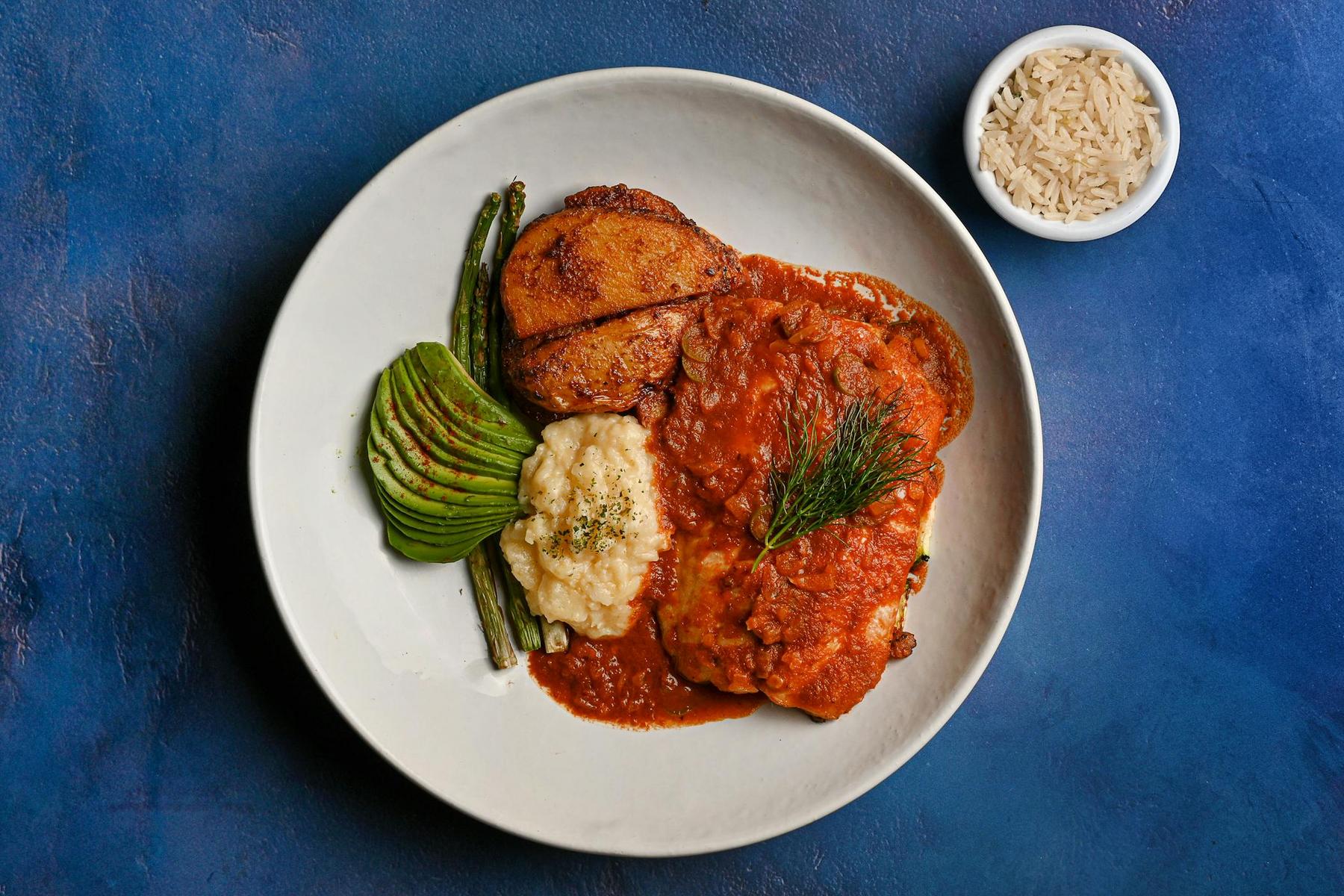 A plate with chicken in tomato sauce, mashed potatoes, fried plantain, avocado slices, green beans, and a small bowl of white rice on a blue background.