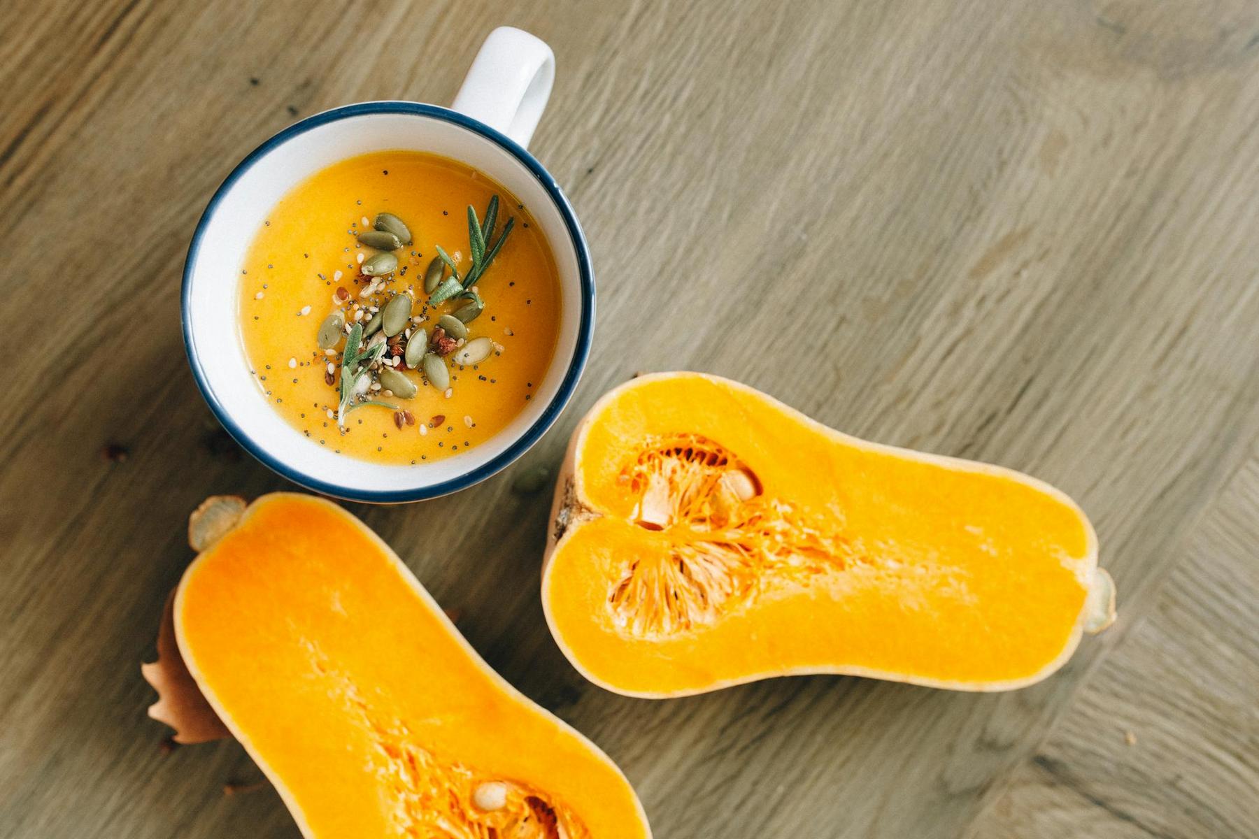 A cup of pumpkin soup garnished with seeds and herbs sits next to a halved butternut squash on a wooden surface.