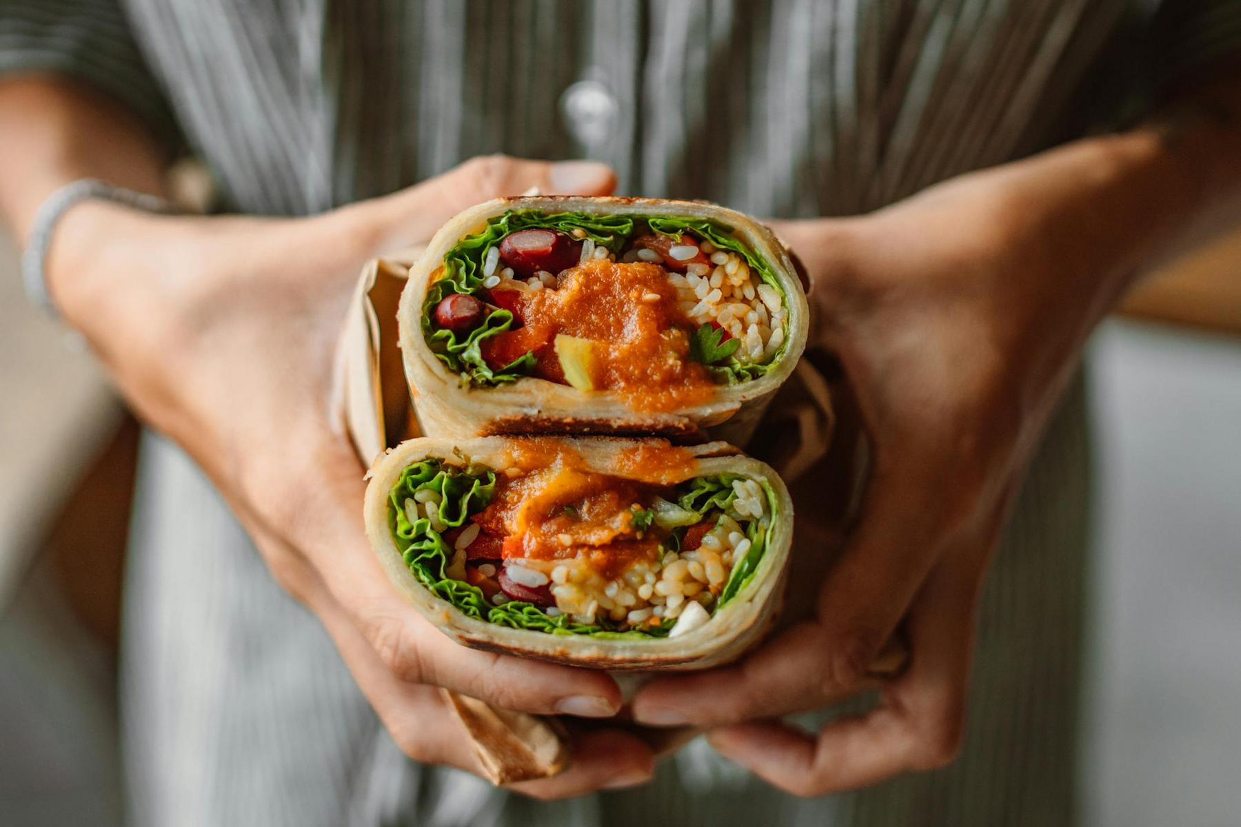 A person holding two halves of a vegetable wrap filled with lettuce, beans, rice, and sauce, with the cross-sections facing the camera.