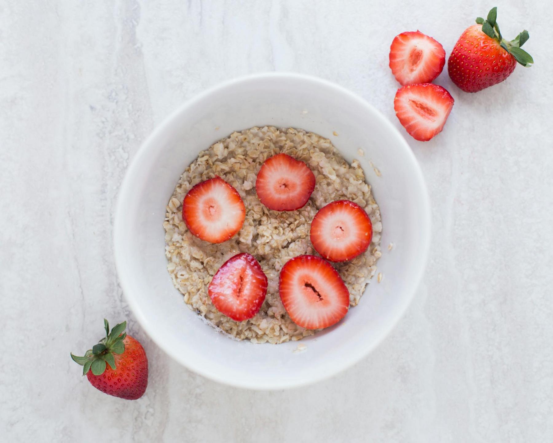 A bowl of oatmeal topped with sliced strawberries, with additional whole and halved strawberries on a light surface beside the bowl.