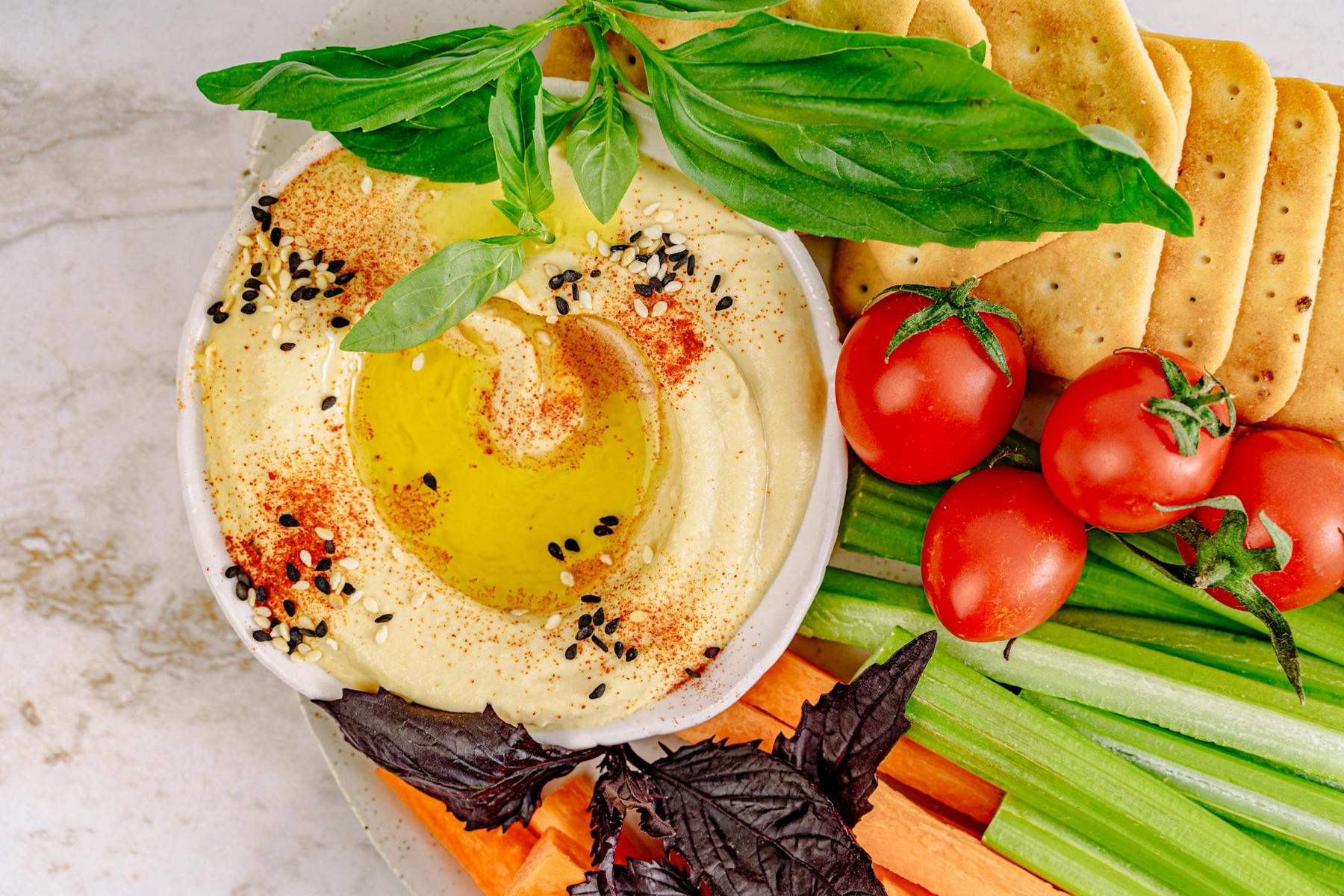 A bowl of hummus topped with olive oil, herbs, and spices, surrounded by crackers, cherry tomatoes, celery, carrots, and fresh basil leaves.
