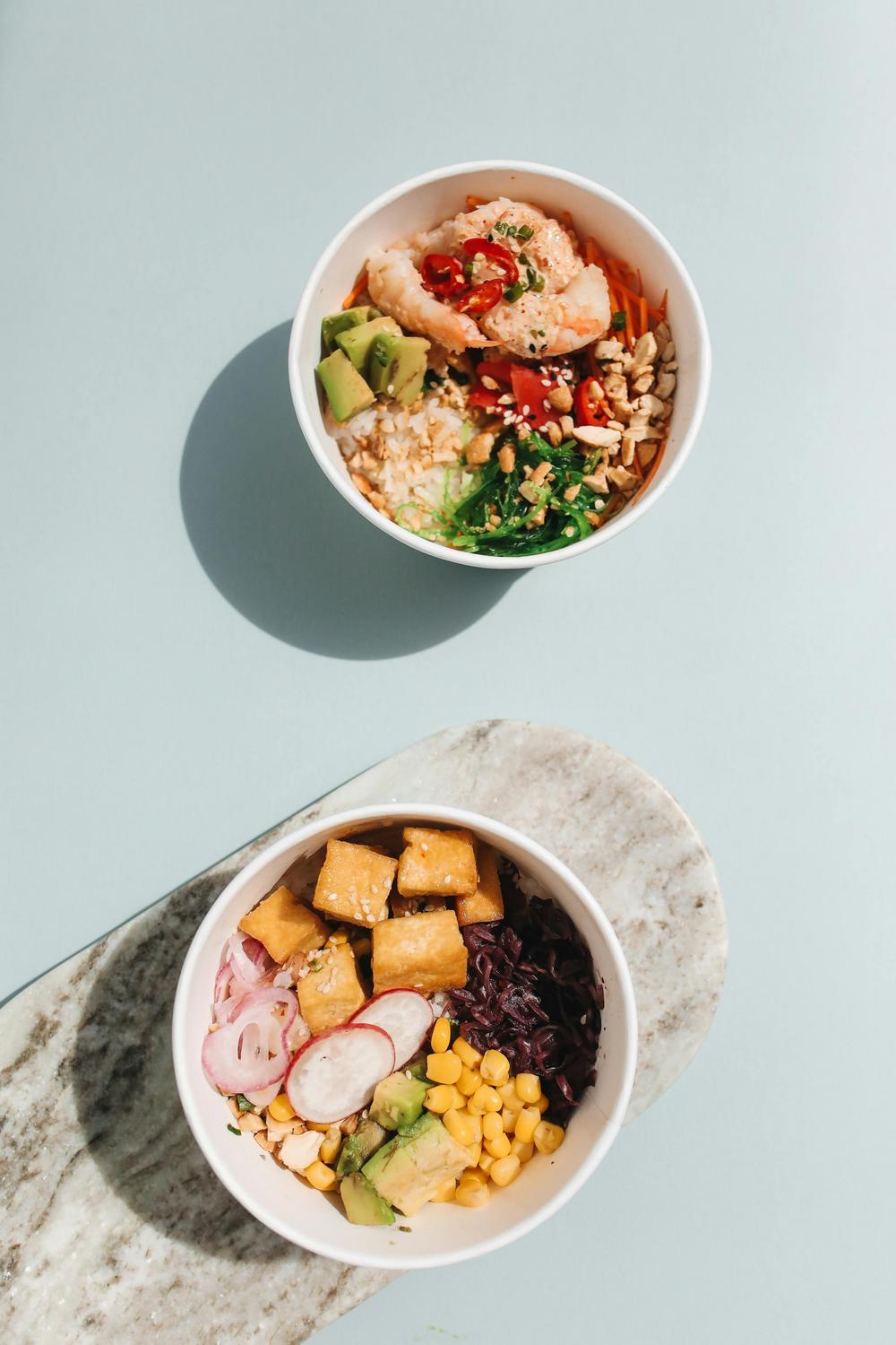 Two bowls of assorted poke salad with ingredients including rice, tofu, avocado, corn, seaweed, radish, shrimp, and peanuts, placed on a light blue surface.