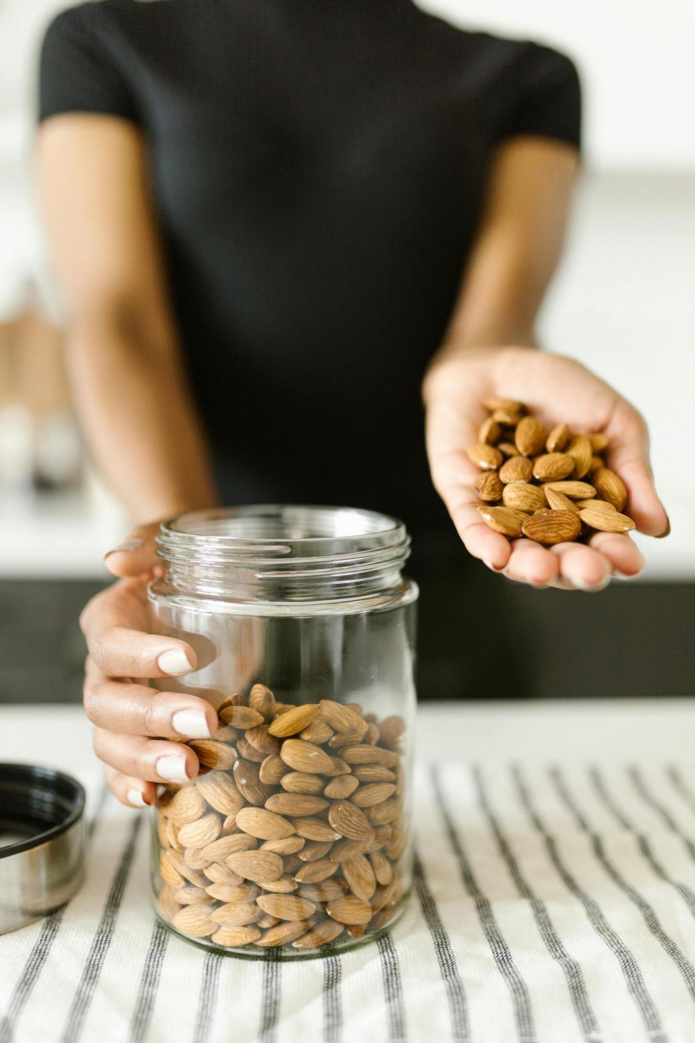 A person holds a handful of almonds above an open glass jar filled with almonds on a striped cloth.