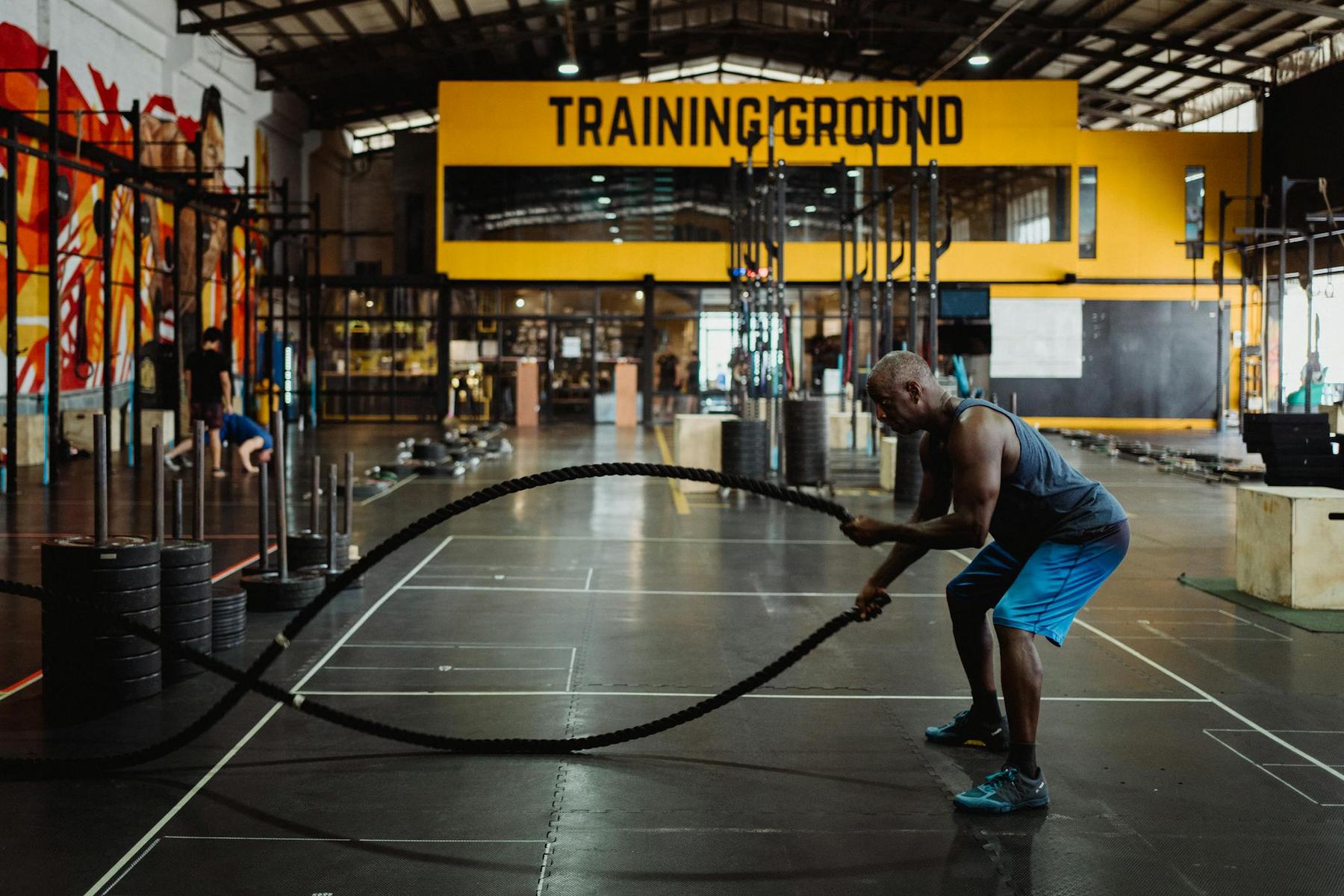 A man exercises with battle ropes in a large gym with "TRAINING GROUND" displayed on a yellow sign in the background.
