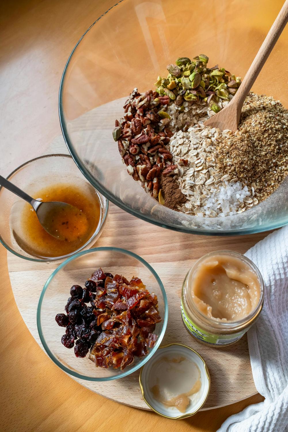 A glass bowl with oats, seeds, nuts, and cinnamon sits next to bowls of dried fruit, sauce, and a jar of nut butter on a wooden surface.