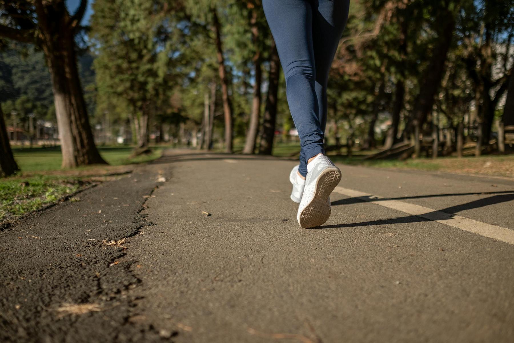 Person wearing white sneakers and blue leggings walking or jogging on a paved path in a park with trees on both sides.