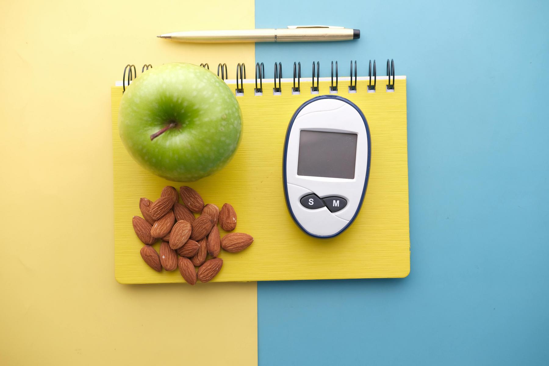 A green apple, almonds, a glucose meter, and a pen rest on a yellow spiral notebook, placed on a two-tone blue and yellow background.