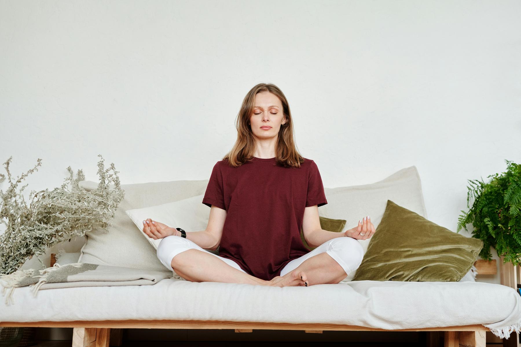 A woman sits cross-legged on a couch with eyes closed, meditating. She wears a maroon shirt and white pants. Cushions and plants are arranged around her.