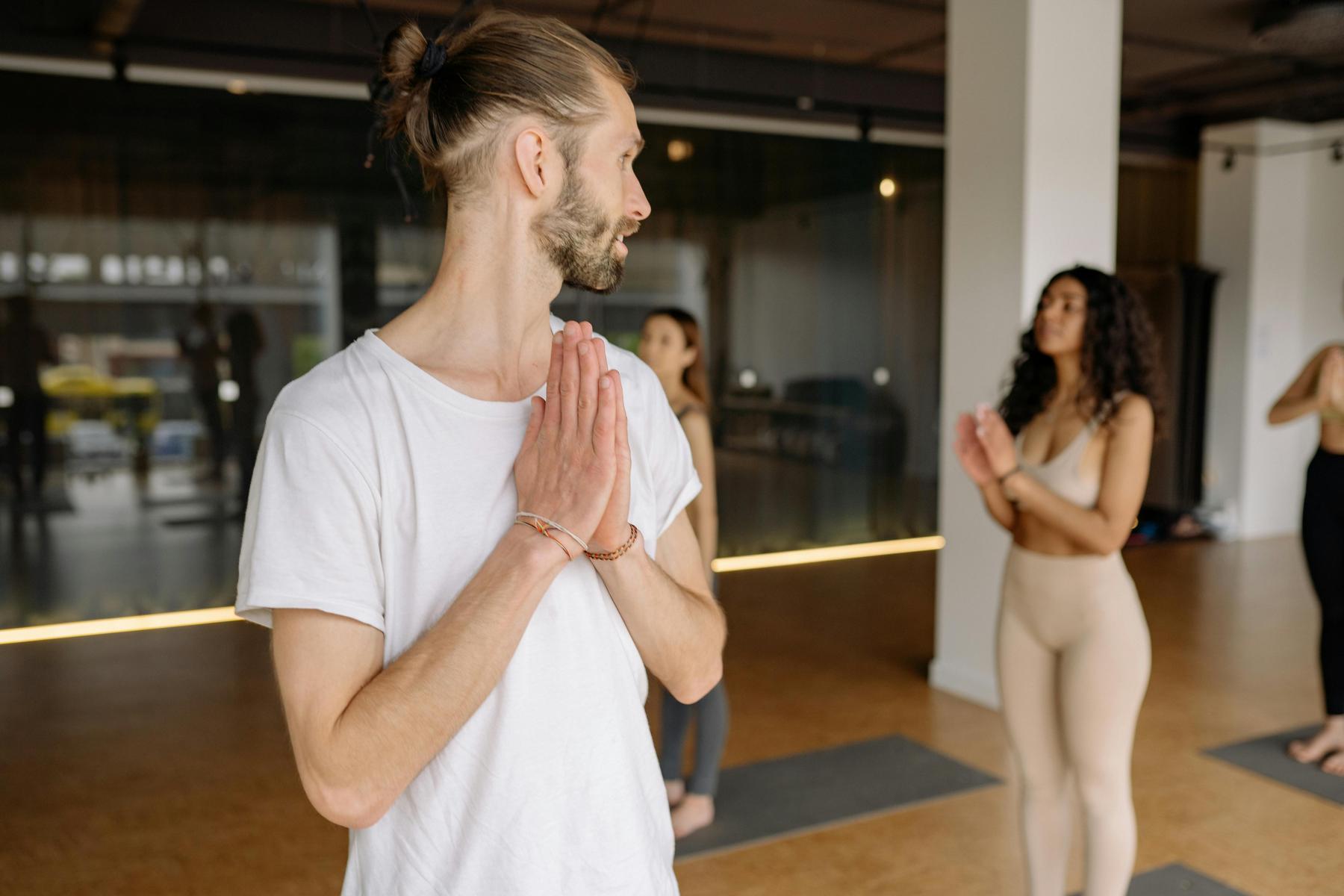 A man in a white t-shirt stands on a yoga mat with hands in prayer position, leading a group yoga class in a studio.