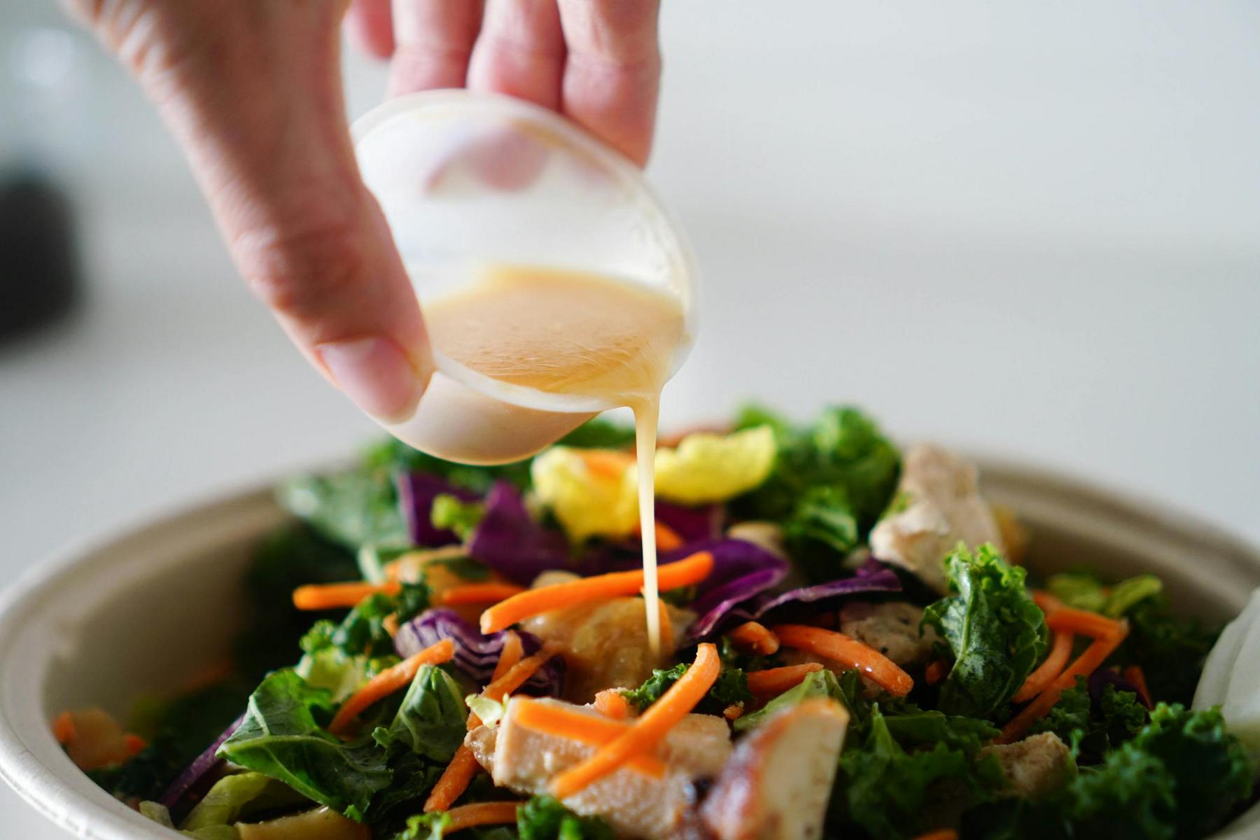 A hand pours dressing from a small cup onto a salad with leafy greens, carrots, and other vegetables in a bowl.