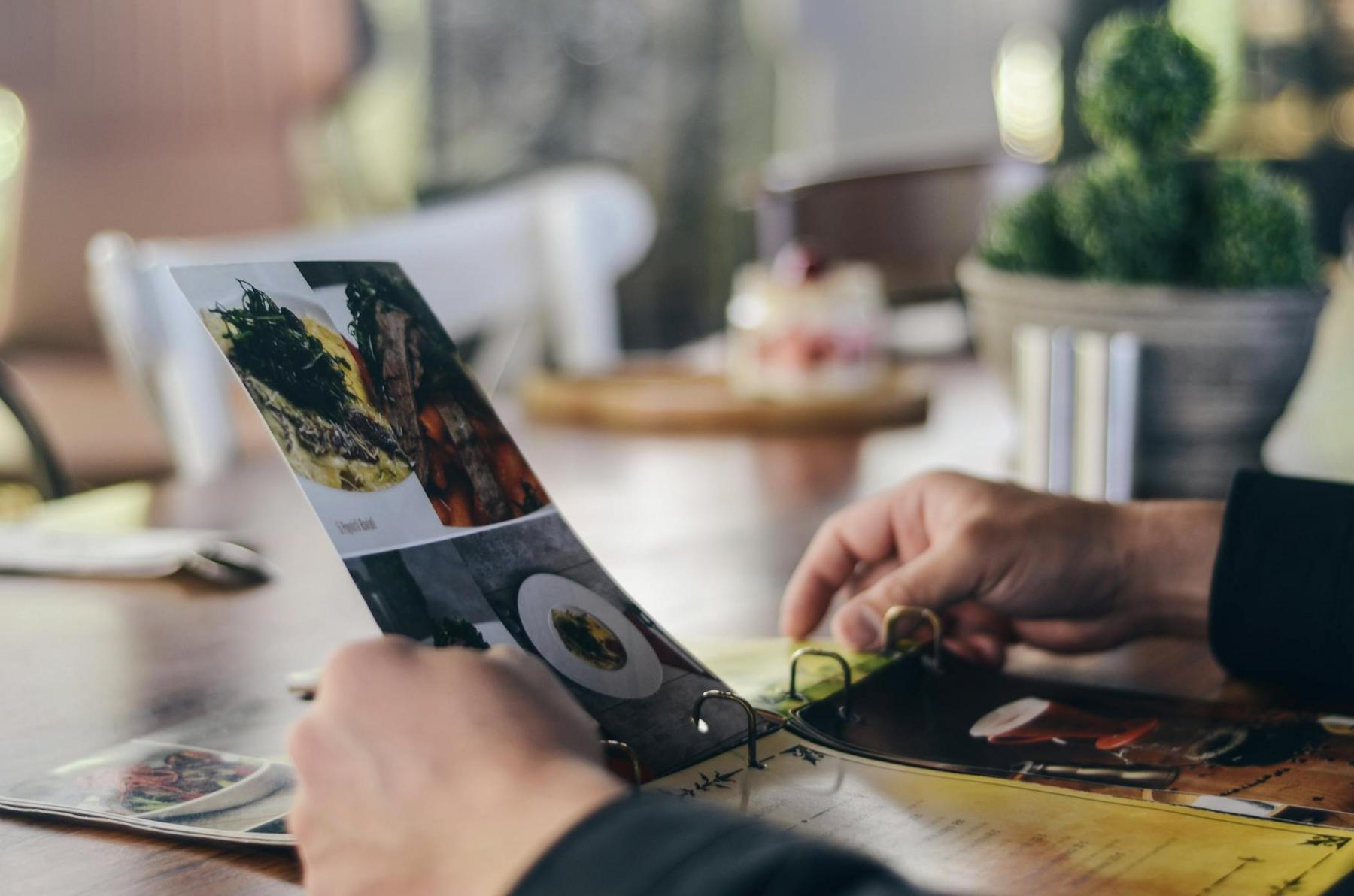 Person browsing a menu at a restaurant table, with a plant and dessert in the background.
