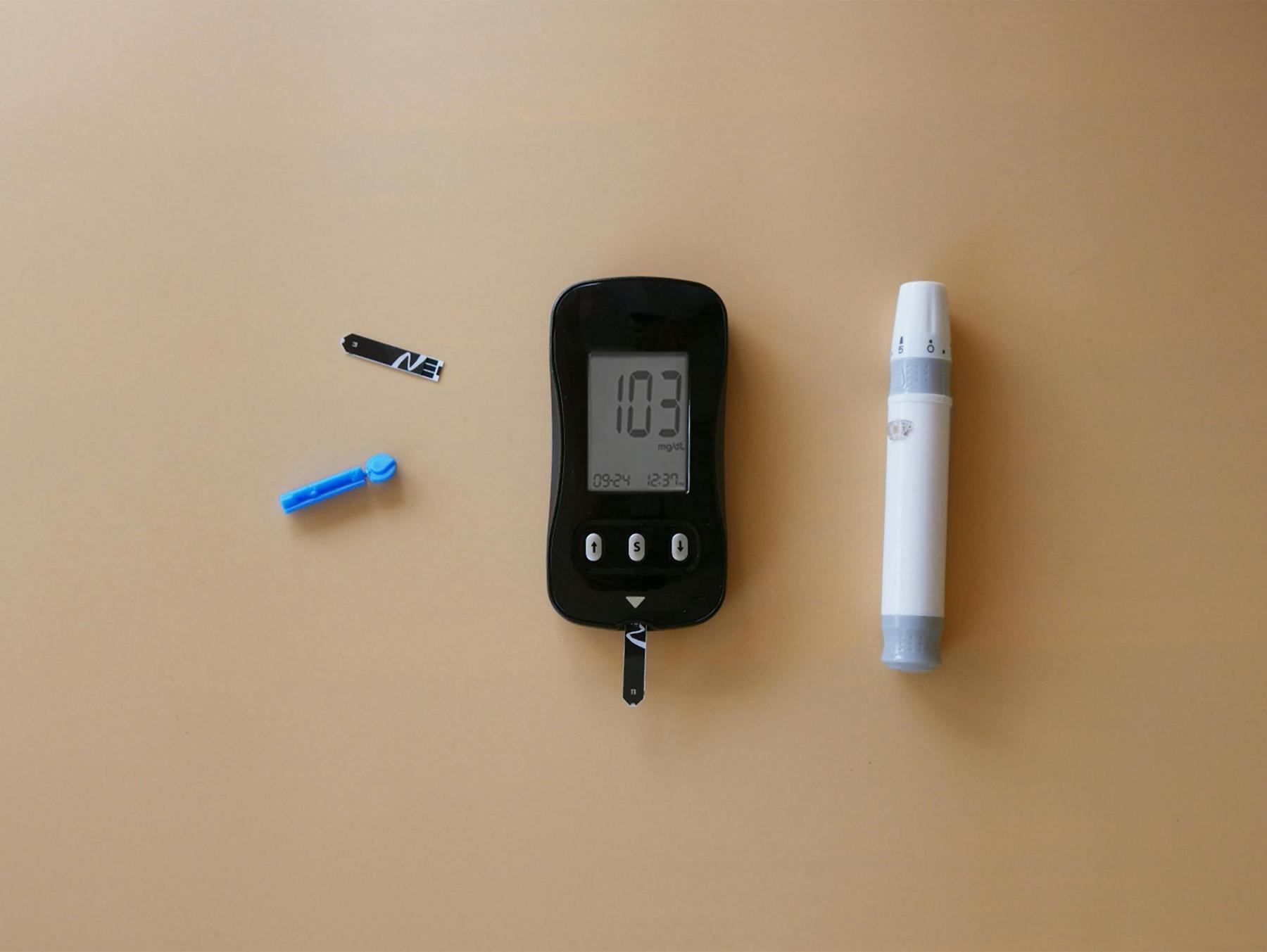A blood glucose meter displays a reading of 103 mg/dL, with a lancet device, test strip, and blue lancet on a beige surface.