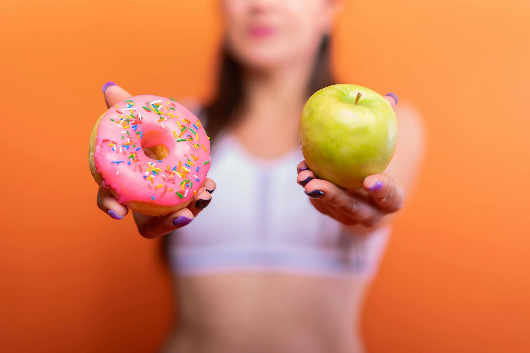 A person in a sports bra holds a pink frosted donut with sprinkles in one hand and a green apple in the other against an orange background.