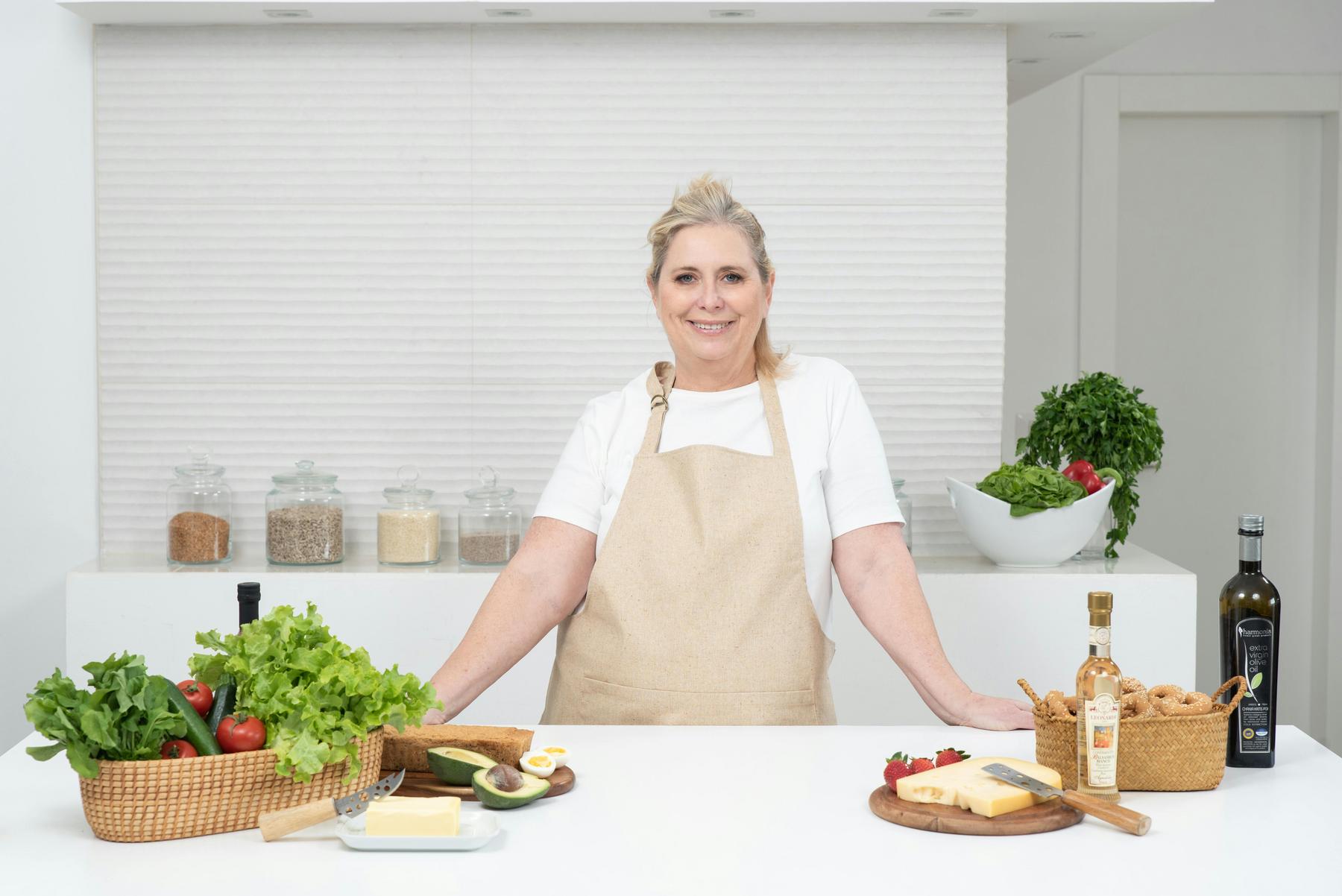 A person in an apron stands in a kitchen, surrounded by various ingredients like vegetables, bread, and oils, with a neutral expression.