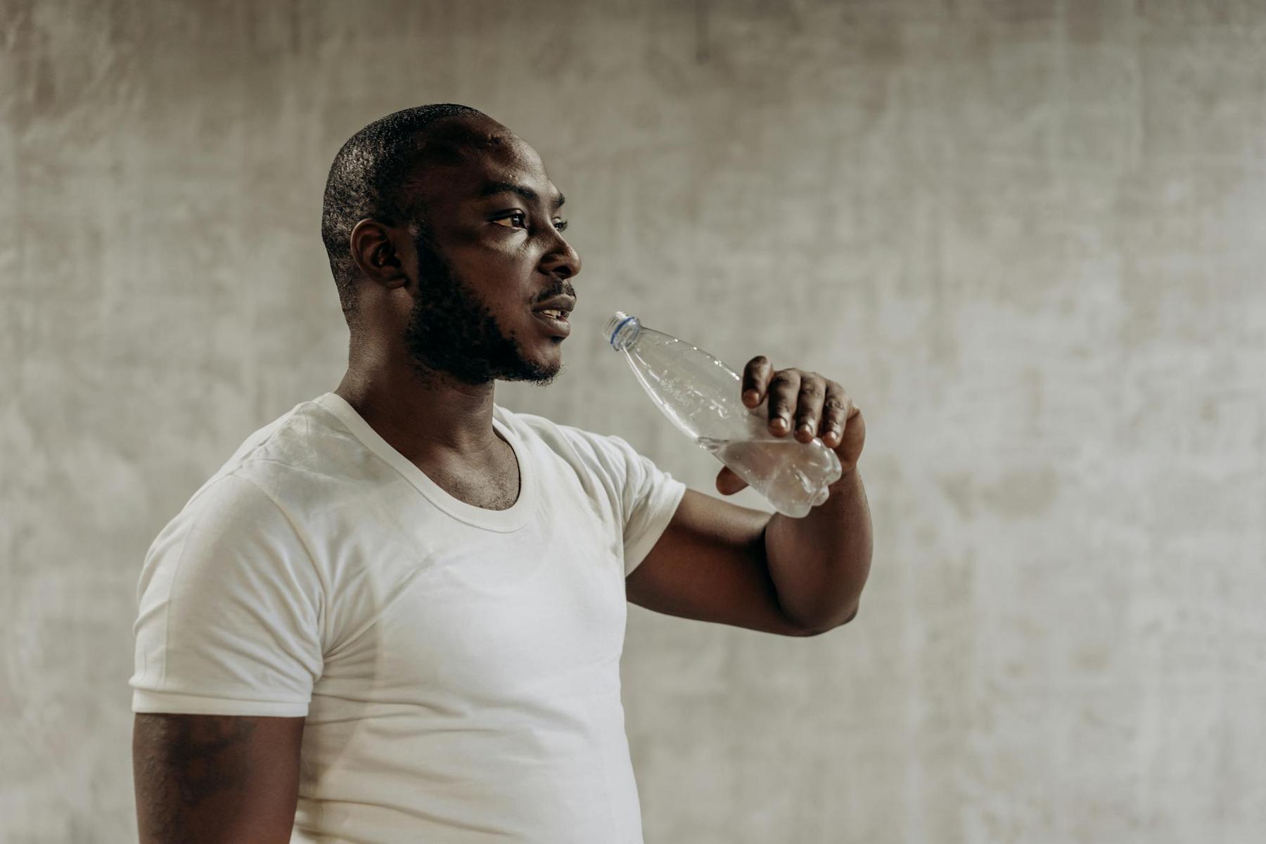 Man in a white t-shirt holding a water bottle, standing against a textured gray wall.