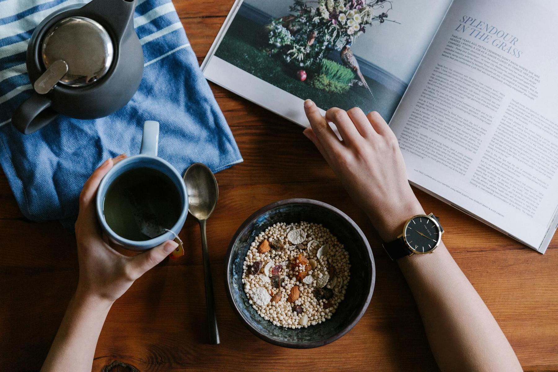 Person reading a magazine at a wooden table with a bowl of cereal, a mug, a spoon, a teapot, and a blue cloth.