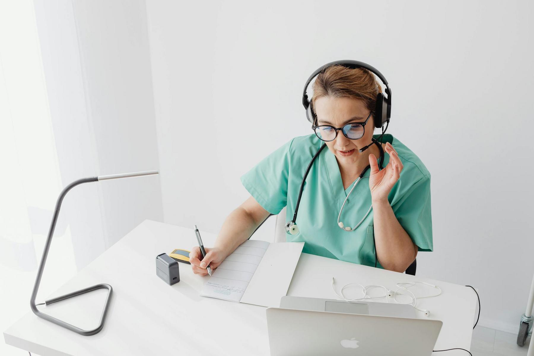 A healthcare professional in scrubs and a headset writes notes while speaking during a video call at a desk with a laptop and medical equipment.