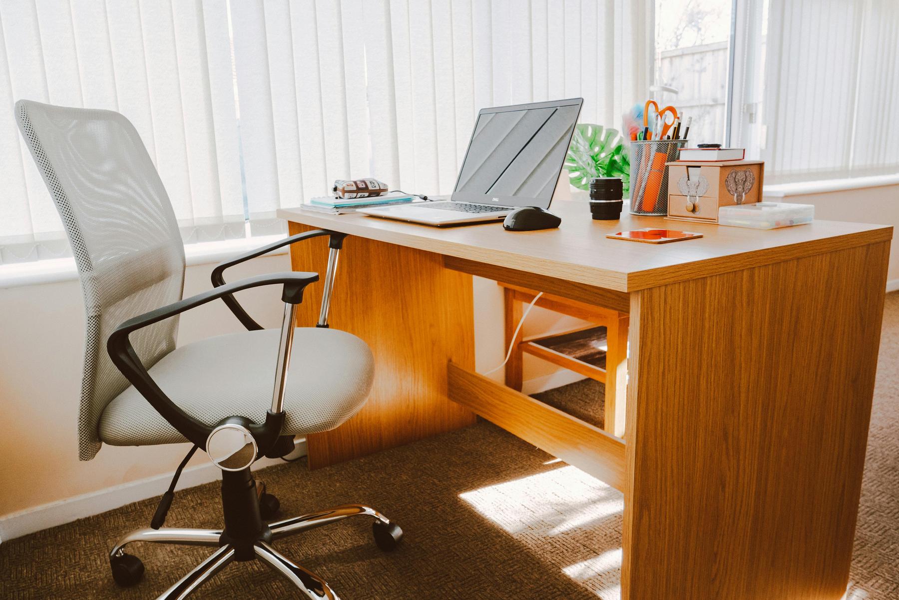 A wooden desk with a laptop, pens, a potted plant, and other office items. An office chair is positioned in front of the desk. Vertical blinds cover the sunny window behind.