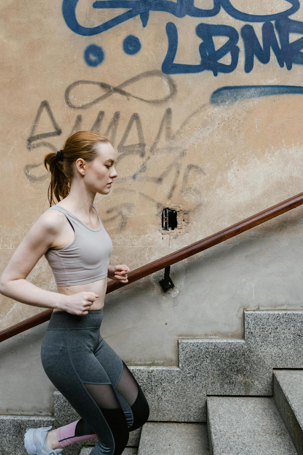 A woman in athletic wear jogs up outdoor concrete stairs next to a wall with graffiti.