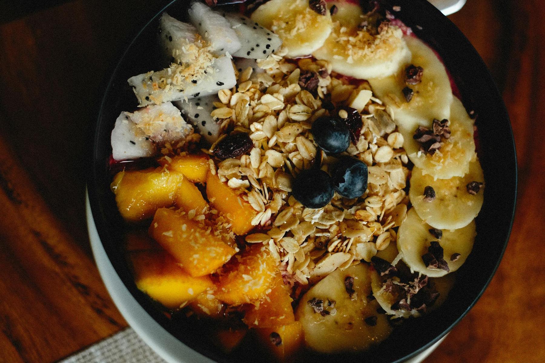 A bowl of granola topped with banana slices, mango chunks, dragon fruit pieces, blueberries, and shredded coconut on a wooden table.