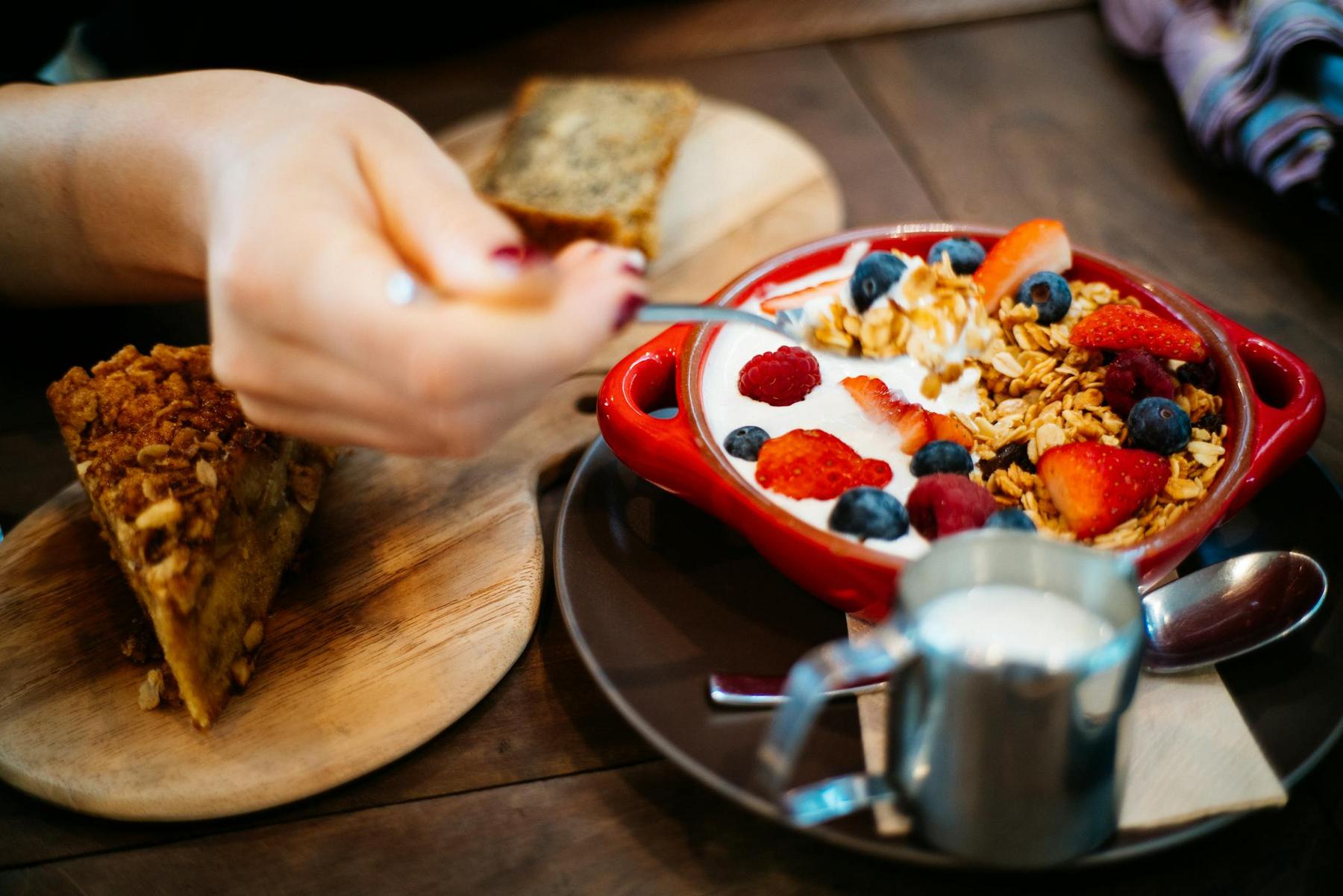 A person holds a spoon over a bowl of yogurt topped with granola, strawberries, blueberries, and raspberries; a slice of cake and cream are nearby on a wooden table.