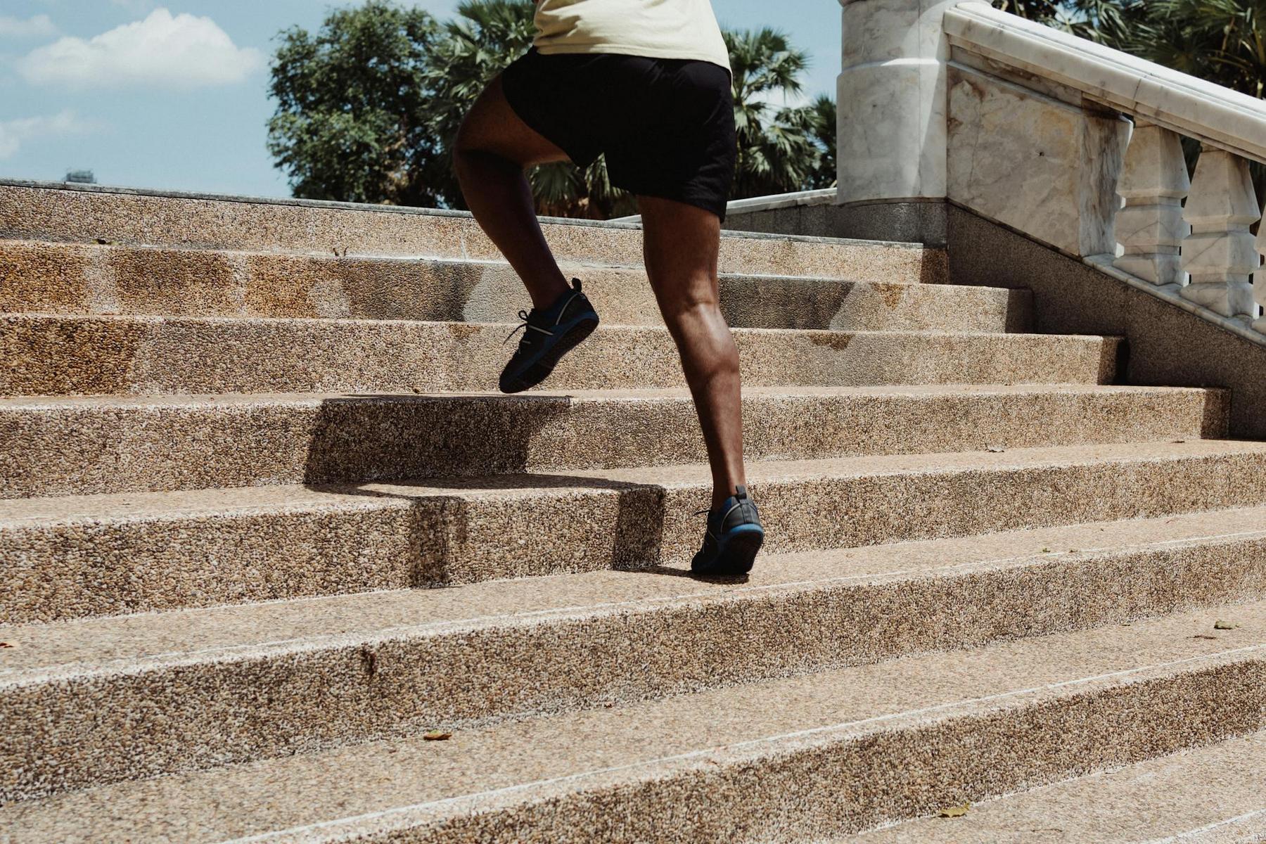 Person in athletic wear running up outdoor stone steps beside a white railing, with trees and a blue sky in the background.