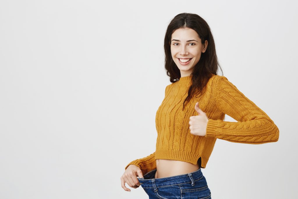 A woman in a mustard sweater gives a thumbs up and smiles, proudly holding out the waistband of her jeans, showcasing her weight loss journey.