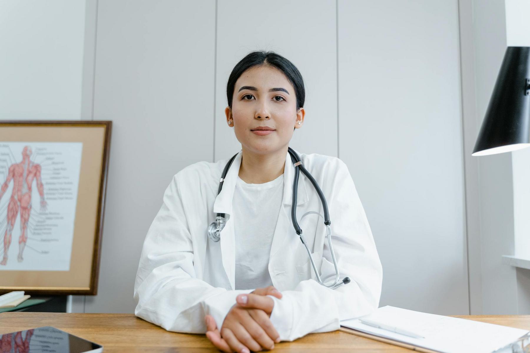 A person in a white coat with a stethoscope sits at a desk with folded hands. A framed anatomy chart and lamp are in the background.