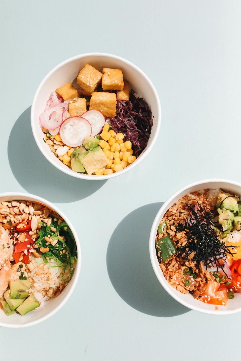 Three white bowls on a light surface, each filled with assorted ingredients including tofu, corn, radish, avocado, rice, seaweed, and vegetables.