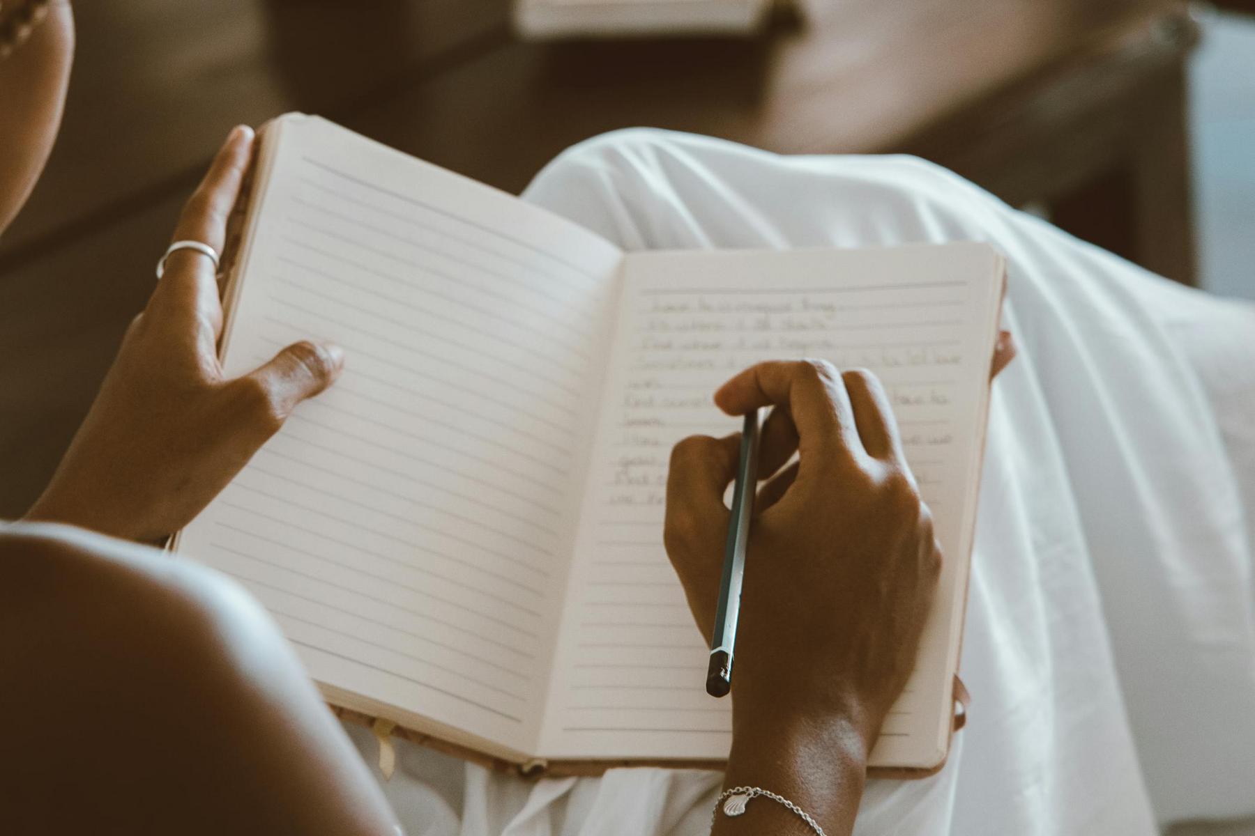 A person writes in a lined notebook with a pen while sitting, wearing a white garment and a bracelet on their wrist.