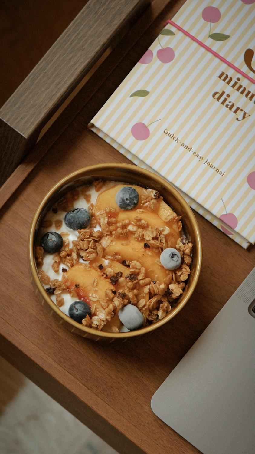 A bowl of yogurt topped with granola, blueberries, and peach slices on a wooden table next to a striped notebook and part of a laptop.