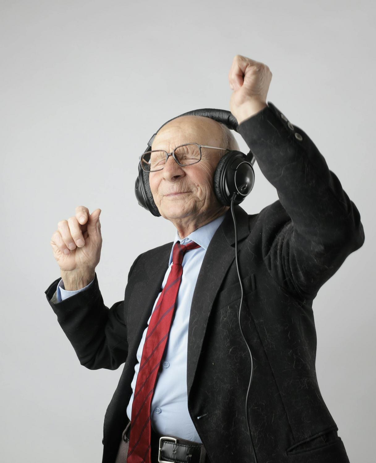 Elderly man in a suit and red tie wearing headphones, eyes closed, and raising his arms as if enjoying music.