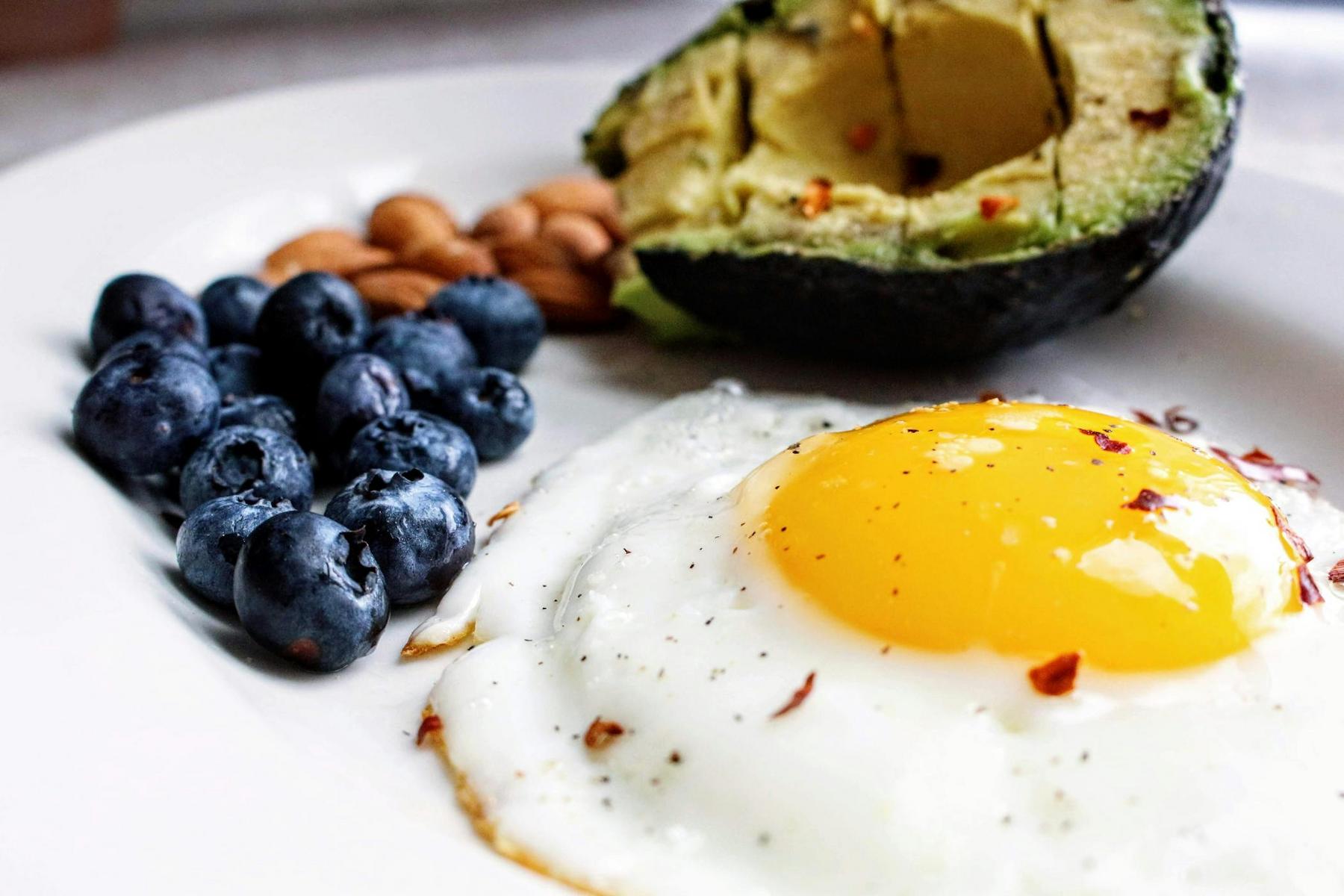 A plate with a sunny-side-up egg, half an avocado with seasoning, a cluster of blueberries, and a handful of almonds.