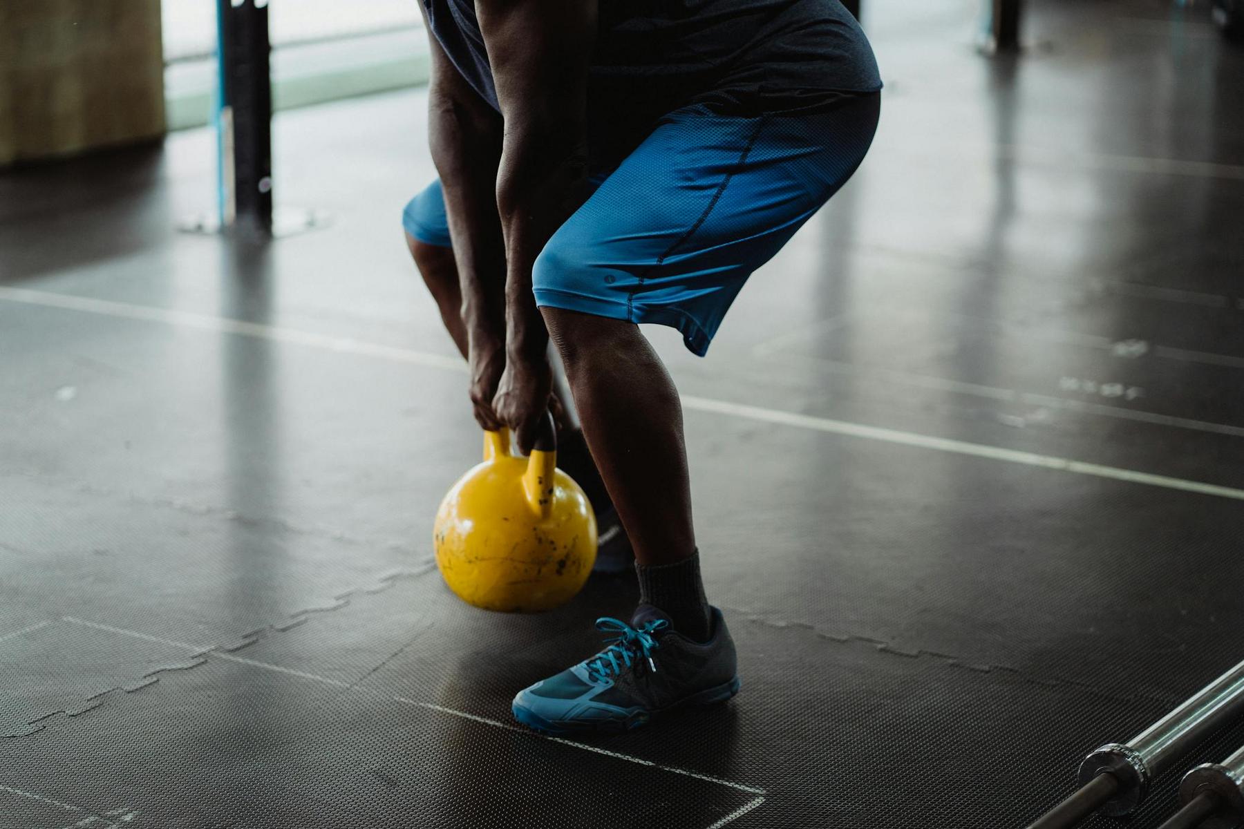 A person wearing blue shorts and sneakers is lifting a yellow kettlebell in a gym setting.