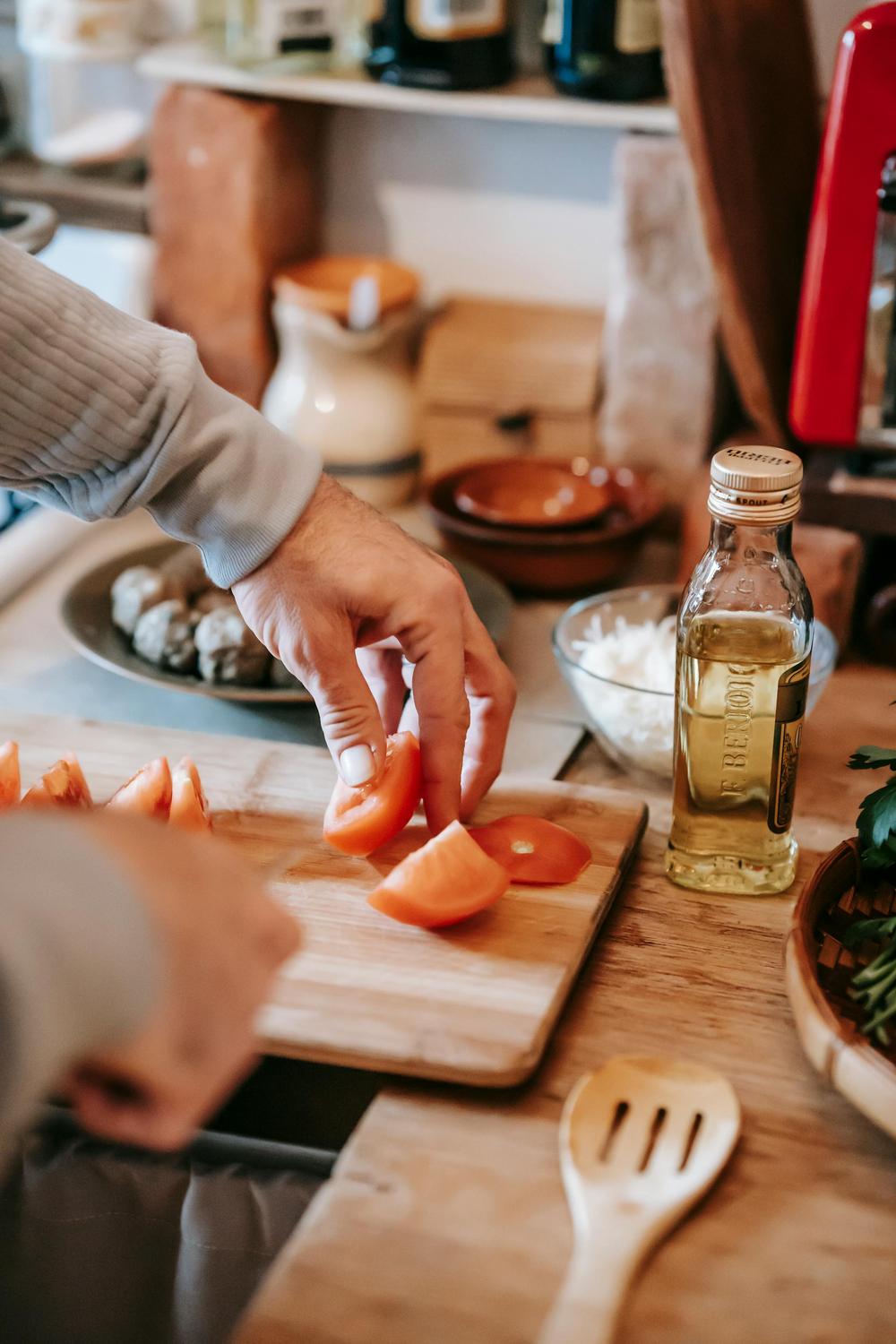 A person slices tomatoes on a wooden cutting board in a kitchen. A bottle of olive oil, a wooden spoon, and various ingredients are visible on the counter.