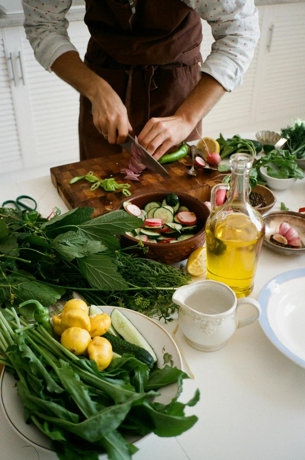 A person slices vegetables on a cutting board surrounded by fresh produce, herbs, a bottle of olive oil, and kitchen utensils on a countertop.