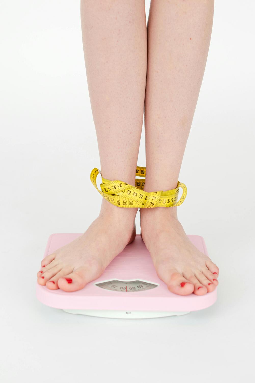 Person standing on a pink bathroom scale with a yellow measuring tape wrapped around their ankles.