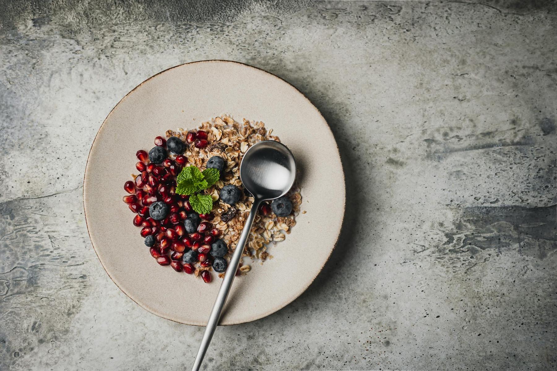 A plate of granola topped with pomegranate seeds, blueberries, and mint, with a metal spoon resting on the plate, on a gray textured surface.
