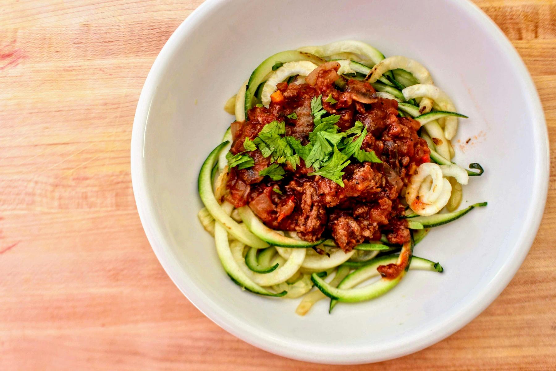 A white bowl contains zucchini noodles topped with tomato-based meat sauce and garnished with fresh cilantro, placed on a wooden surface.